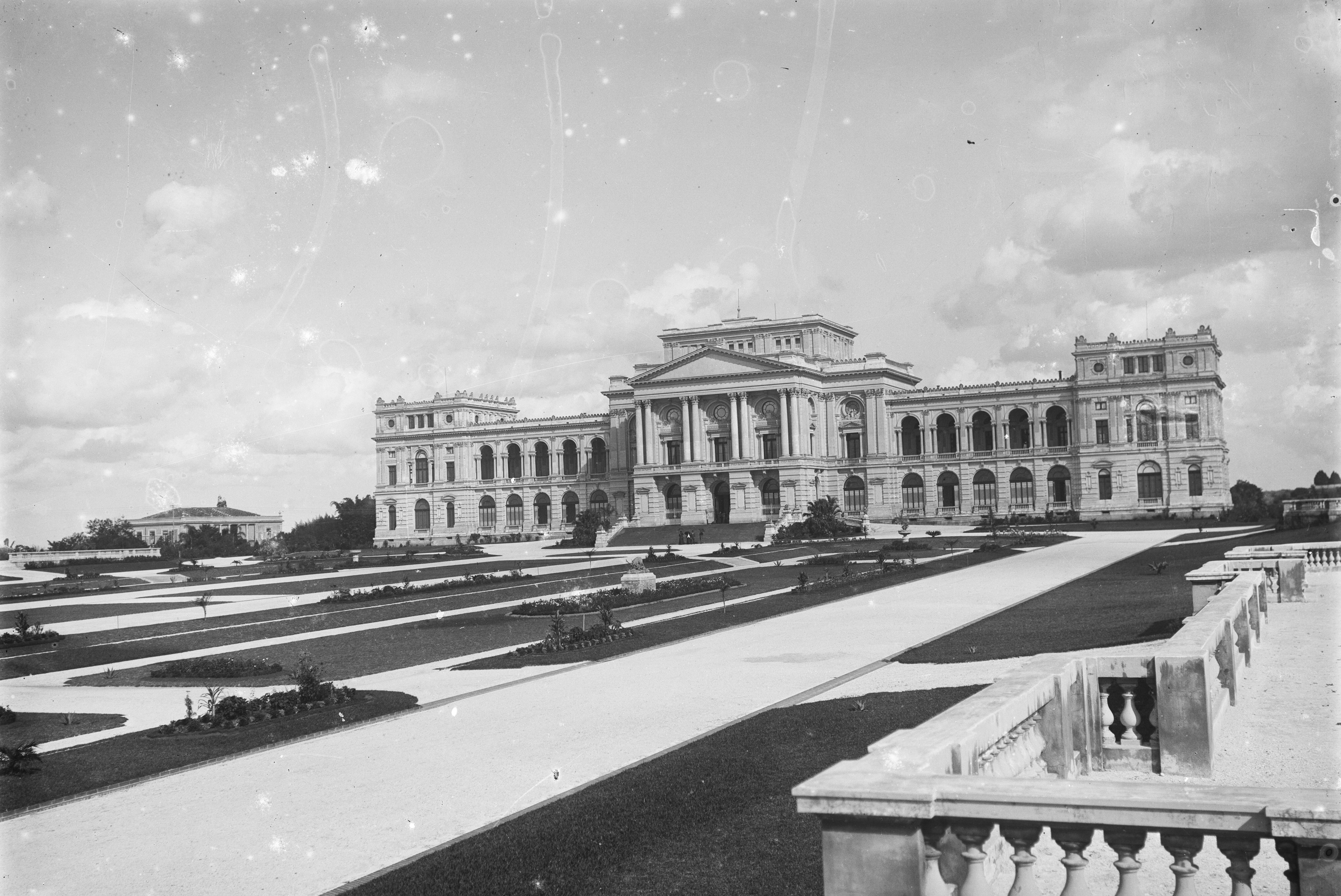 Audiodescrição: Negativo de vidro da fachada e do jardim do Museu do Ipiranga. É uma imagem em preto e branco, no qual vê-se, ao fundo, o imenso edifício, que possui o corpo composto por três torres interligadas por galerias. À frente há um imenso jardim que mistura gramados em várias áreas retangulares, diferentes tipos de árvore, flores vermelhas e alguns postes de luz. Ao fundo há um céu repleto de nuvens.