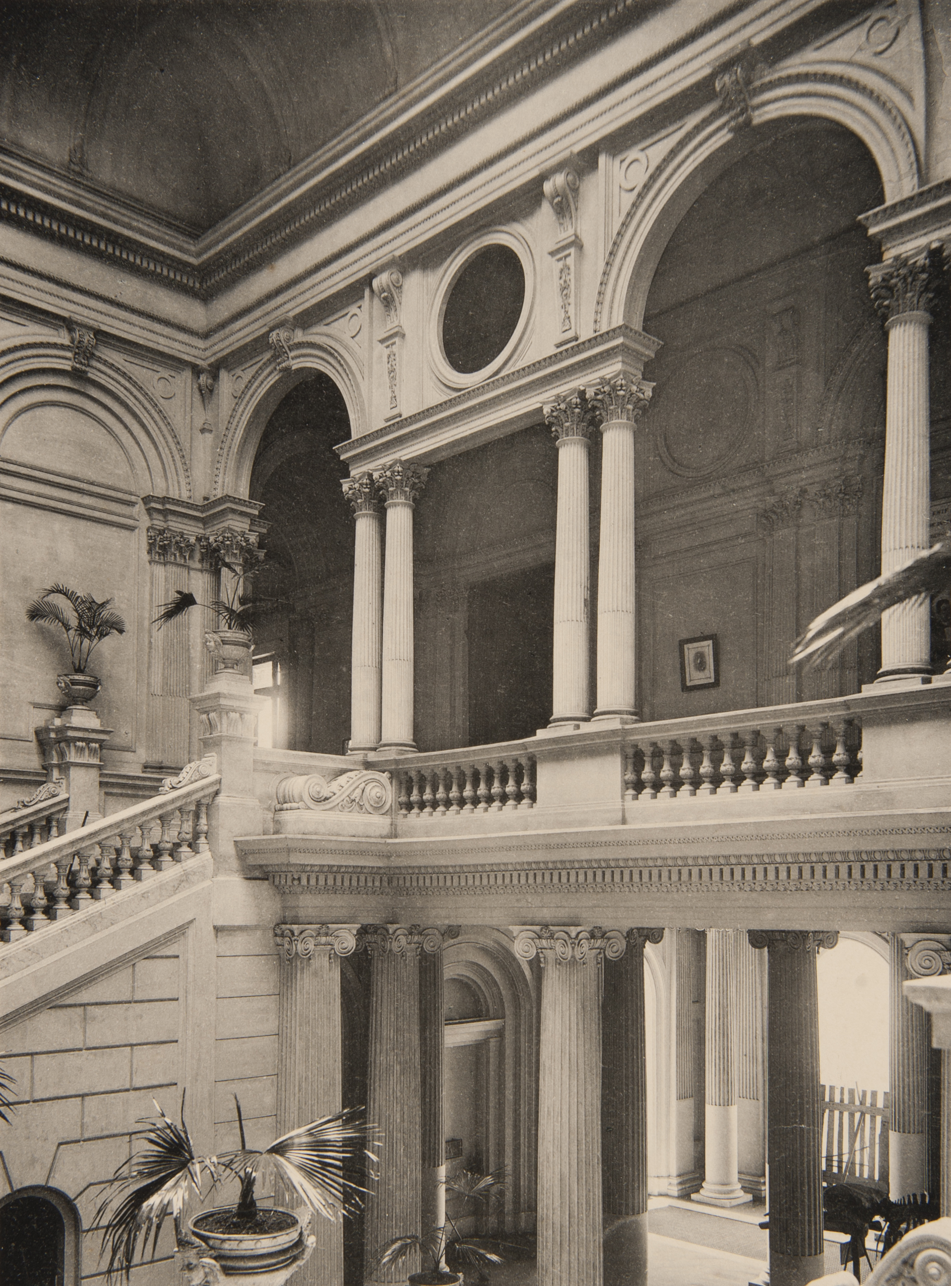 Audiodescrição: Foto em tons amarronzados de dois andares do museu, vistos da escadaria central. No andar inferior há vasos com plantas no chão e diversas colunas de sustentação. No andar superior há, à esquerda, parte da escadaria que dá acesso a um imenso corredor, onde há um corrimão formado por balaústres e, sobre este corrimão, há áreas com o pé direito alto, arqueadas, sustentadas por colunas duplas. No final da escadaria, sobre colunas baixas, há vasos com plantas.