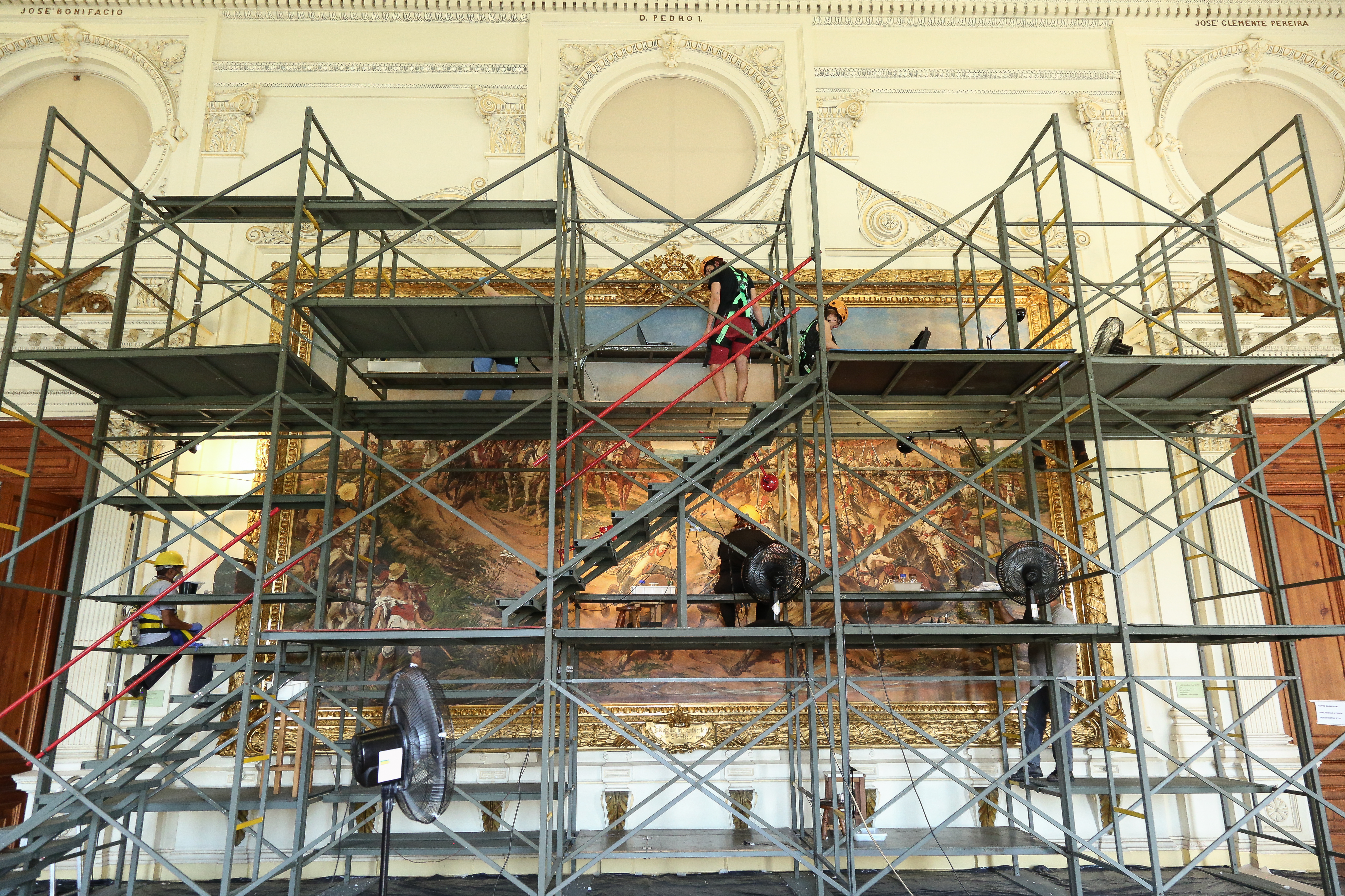 Audiodescrição: Foto da obra Independência ou Morte. À frente dela há um imenso andaime de três andares. Algumas pessoas estão nos três andaimes, virados de costas, tocando na obra.