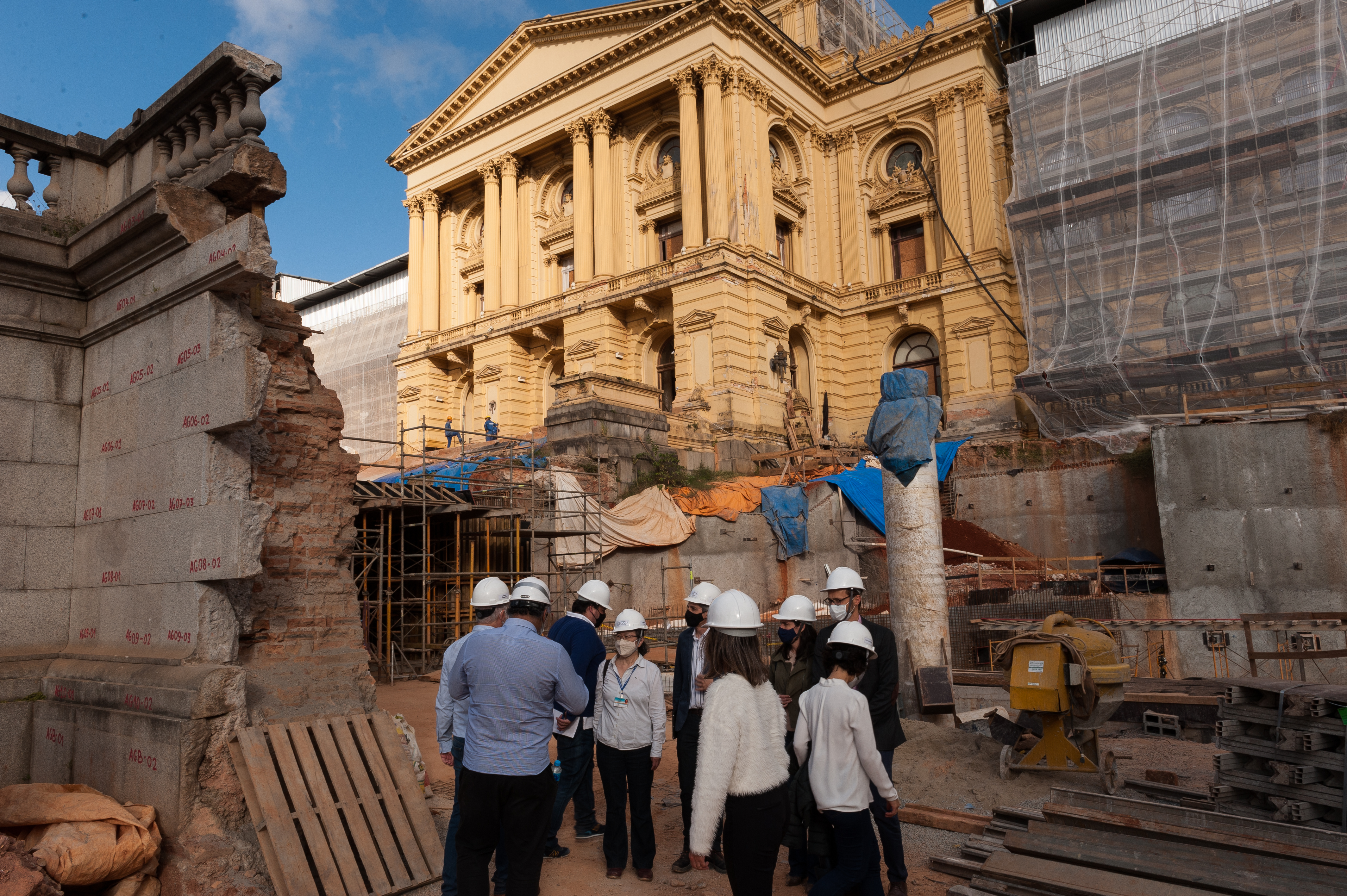 Audiodescrição: Foto do Museu do Ipiranga em obras. O imenso edifício amarelo está com as galerias cobertas por redinhas de proteção. À frente há andaimes, equipamentos e um grupo de pessoas com capacetes brancos. À esquerda, numa outra construção, vê-se uma parede demolida.