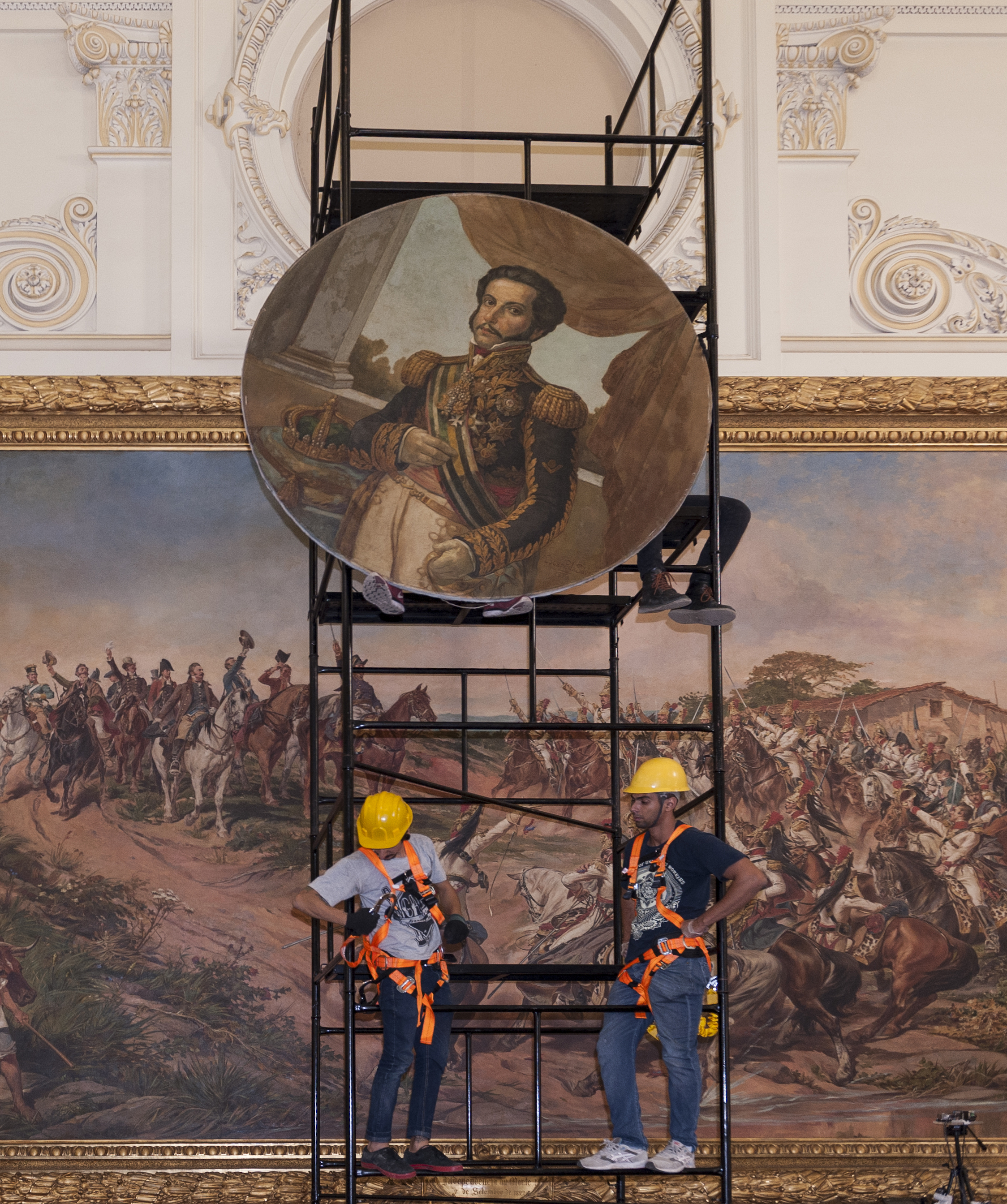 Audiodescrição: Foto de um grande salão. Em frente à pintura “Independência ou Morte há um andaime. Acima da pintura há espaços circulares, nos quais estão as pinturas de grandes personalidades. A pintura de Dom Pedro Primeiro, em moldura circular, está apoiada nos pés de uma pessoa que está sentada no alto do andaime, totalmente coberta pela pintura. Ao lado dela se veem as pernas de outra pessoa. Abaixo, no primeiro andar do andaime, estão dois homens com capacetes amarelos. Um deles prende um gancho às fitas laranja de segurança que estão presas ao próprio corpo.